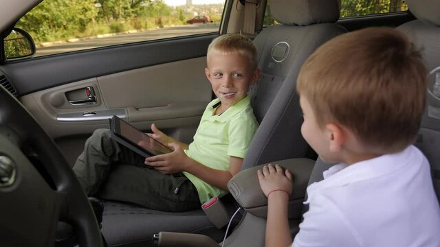 Two cheerful boys in polo shirts are sitting in the front seats of a car and playing video games on a tablet.