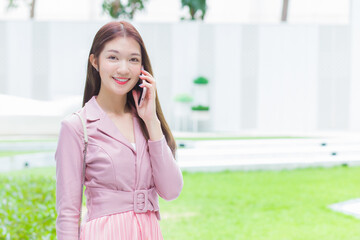 Young Asian business professional in pink dress look at camera is calling phone seriously with someone at office building.