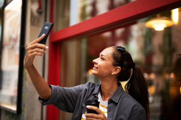 Happy girl enjoying in sunny weather and fresh coffee. Gorgeous casual brunette drinking coffee to go. Young woman having video call.