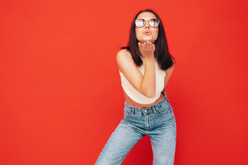 Portrait of young beautiful smiling female in trendy summer clothes. Sexy carefree woman posing near red wall in studio. Positive model having fun indoors. In sunglasses. Gives air kiss