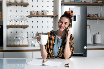 Young businesswoman drinking coffee and talking on the smartphone at the kitchen. Creative female business owner planning and contracts a job over the phone at home in the morning.