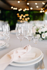 Wedding dining table. Side view of white round plates and glasses close up on background of lights