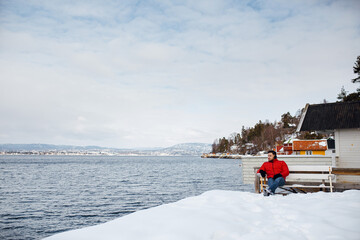 Young traveler man in red winter jacket sitting on the porch of the traditional norwegian rorbu house near the sea