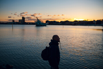Traveler man with backpack standing on the sea coast and looking at the bay at sunset. Dusk