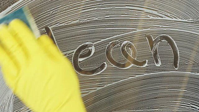 Soapy glass surface with inscription Clean. Hand in yellow rubber glove washes away the word Clean.