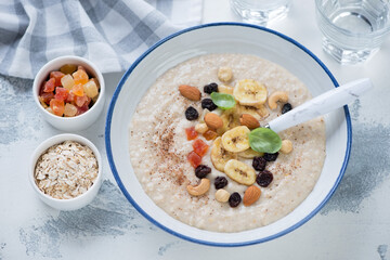 Plate of oatmeal porridge topped with dried banana, candied fruits and nuts, horizontal shot on a white concrete background