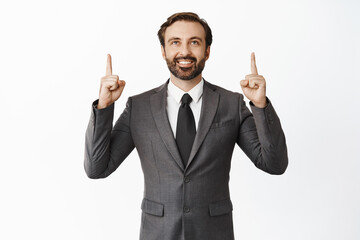 Happy businessman in suit pointing and looking up with pleased face, showing advertisement, standing over white background