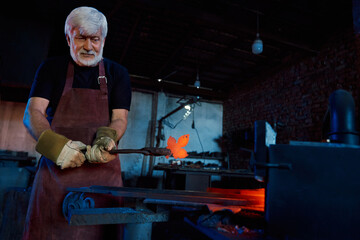 Grey haired man in protective apron and gloves working at forge with heated metal. Experienced...