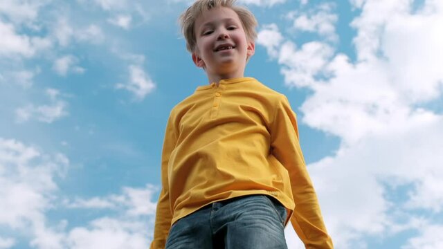 Happy child jumps very high on a trampoline against the blue sky. Blond boy jumps on a trampoline. Slow motion bottom view of a cheerful boy jumping on a trampoline. Children happiness.