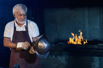 Skillful aged craftsman in protective apron and gloves making gladiator helmet from steel at forge....