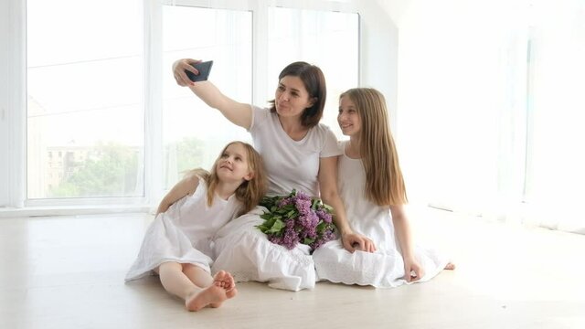 Mother with daughters and lilac bouquet making selfie with smartphone together. Children with parent and flowers doing photos