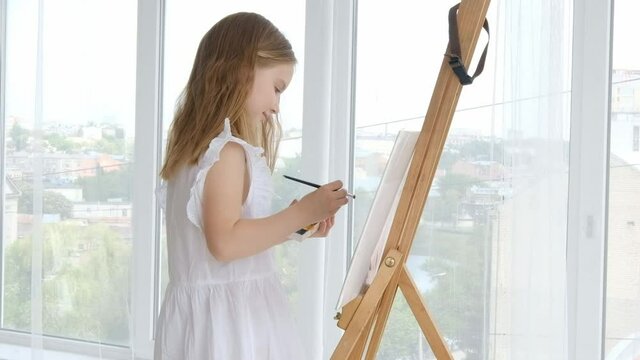 Preteen girl with easel drawing at canvas and holding color palette paints. Cute female kid with paintbrush develop picture at home