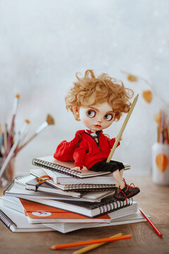 A puppet boy sits on the artist's desk with a pencil in his hand on a stack of sketchbooks