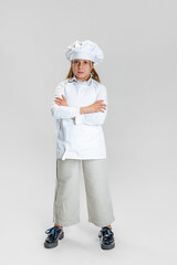 Portrait of little cute girl in white cook uniform and huge chef's hat standing with arms crossed isolated on white studio background.