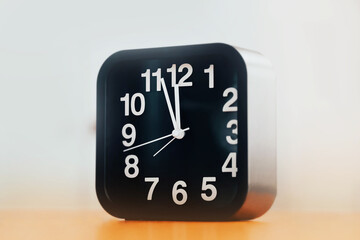 A black modern clock stands on a wooden table and shows five minutes to twelve. Wake up late in the morning. Lunch time. Five minutes until the New Year.