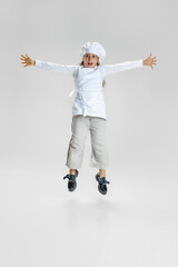 Dynamic image of little cute girl in white cook uniform and huge chef's hat jumping isolated on white studio background.