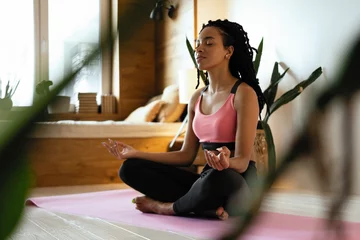 Abwaschbare Fototapete Young black woman doing yoga at home in the lotus position. © opolja