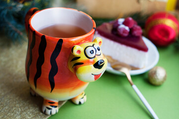 Mug in the shape of a tiger with tea and raspberry cheesecake on a shining gold and green background. Cup in the form of a tiger - a symbol of 2022. New year tea and dessert with christmas decorations