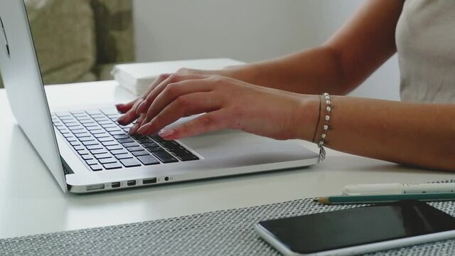 Female hands type on laptop keyboard touching touchpad with fingers smart phone lying near close up. No face woman sits on sofa freelancing working at home writes post comments in social networks