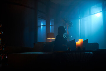 Silhouette of young woman sitting on couch at dark atmosphere and looking on full moon through...