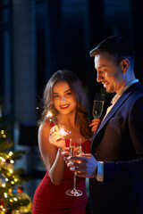 Happy young woman and man celebrating new year with sparklers and champagne. Beautiful couple in stylish clothes staying at home during winter holidays.
