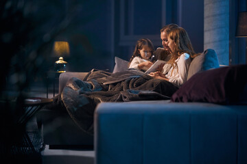 Caring caucasian mother sitting on couch with two pretty daughters under soft blanket and reading...