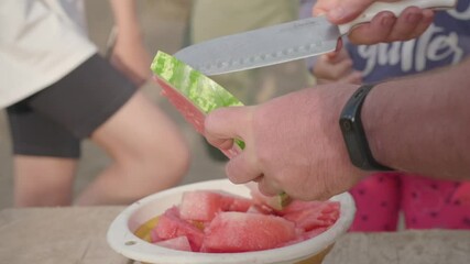 A man cuts a watermelon with a large knife into a plate. Children are walking in the background.