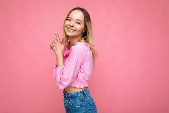 Photo of young positive happy smiling attractive blonde woman with sincere emotions wearing pink blouse isolated on pink background with empty space and making shooting hand gun