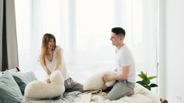 Handsome man and woman wearing pajamas have fun at home. Beautiful couple fights with pillows in the in the bed and smiling. Young wife and husband in the morning time. Concept of happy relationships