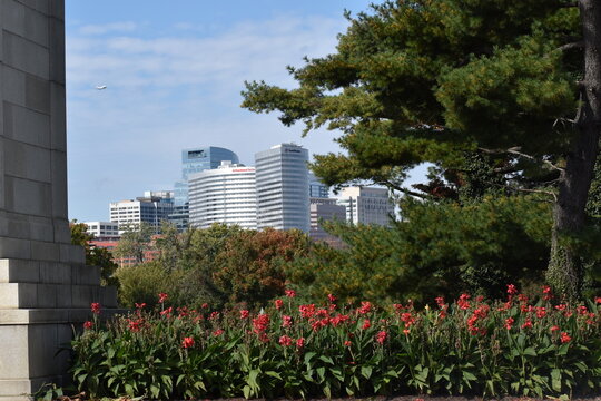 Arlington, Virginia, USA - October 25, 2021: Arlington Skyline as Seen from Memorial Drive with the City Framed by Red Canna  Lilies, an Evergreen Tree and a Concrete Pedestal in the Foreground, and a