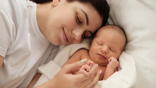 Pretty mother lying in the bed and looking at sleeping newborn baby. Adorable infant child napping and his mom strocking his hand. Girl resting with cute little kid and smiling