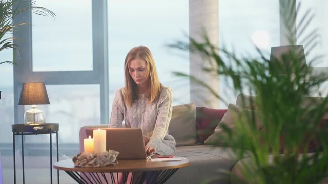 Young caucasian woman with blond hair using wireless laptop while sitting on comfy couch. Attractive female in casual wear surfing internet during free time at home. 