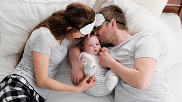Beautiful family wearing same pajamas and sleeping eye masks lying in the bed with little child boy. Young parents kiss their toddler baby in sunny bedroom