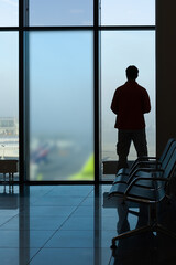 Fototapeta na wymiar Airport Lounge Window. Empty seats and the silhouette of a man looking through the glass at the planes and the take-off field.