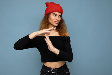 Young emotional dissatisfied pretty brunette woman wavy-haired with sincere emotions wearing black crop top and red hat isolated on blue background with free space and showing pause sign