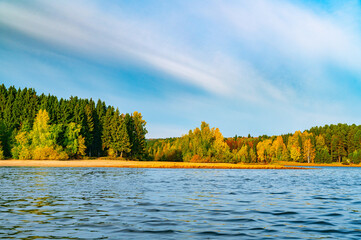 Fototapeta na wymiar Panoramic view of the bright colorful trees along the bank of the Kama River Russia Perm Krai. Autumn colored trees and blue sky, reflected in the calm and shiny water.