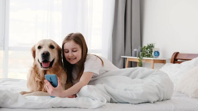 Beautiful preteen little girl sitting in the bed with golden retriever dog and making selfie with smartphone. Cute smiling kid doing photos with pet doggy on cell phone. Child and technologies