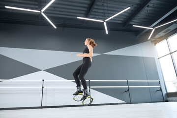 Fototapeta na wymiar Young caucasian girl wearing black sportswear practicing dance moves while doing kangoo jumps. Side view of athletic teenager doing dance cardio exercise in hall, hi tech interior. Sport concept.