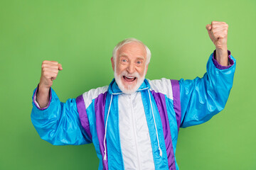 Photo of lucky funky senior man wear sportive suit rising fists smiling isolated green color...