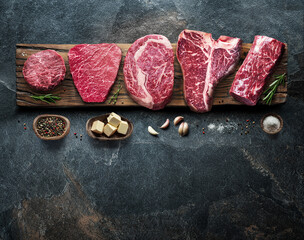 Different raw beef steaks on the wooden board on the grey table. Top view.