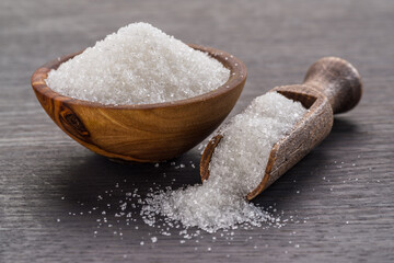 White refined sugar in wooden bowl and spoon on dark wooden table.