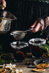 Swedish glögg in glass on dark background. Traditional Nordic drink. Woman pouring mulled wine with spices, orange and raisins.into glasses through sieve.