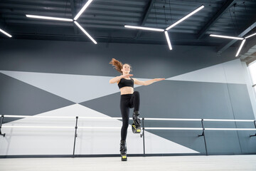 Fototapeta na wymiar Young pretty caucasian girl wearing black sportswear practicing dance moves while doing kangoo jumps and smiling. From below view of sportswoman doing cardio exercise in hall, hi tech interior.