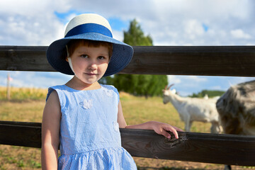 Rural landscape. Portrait of a little girl in a straw hat on the background of a pasture with goats. Selective focus.