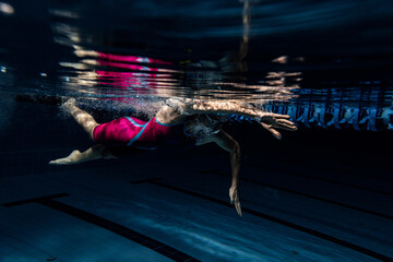 One female swimmer in swimming cap and goggles training at pool, indoors. Underwater view of swimming movements details.