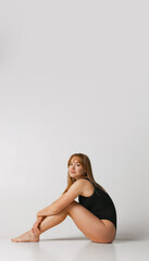 Obraz premium Young beautiful red-headed girl in black swimsuit sitting on floor isolated over gray studio background. Natural beauty concept.