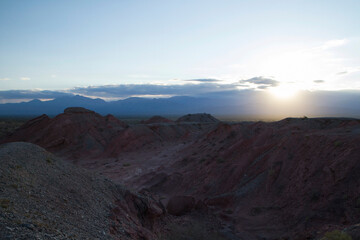 Fototapeta na wymiar The red canyon and valley at sunset. Panorama view of the arid desert, sandstone formations, rocky mountains, lens flare and hiding sun in Talampaya national park in La Rioja, Argentina.