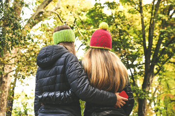 Fototapeta premium View from the back of two girls in the park. Teen walking outdoor in the forest. Young student woman enjoying free time in nature. Women embraced in a beautiful autumn afternoon looks the trees.