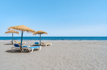 Sombrillas de madera y paja y hamacas sobre la arena de la playa y las aguas del mar mediterráneo un día soleado con cielo azul. Desde Málaga, Costa del sol, Andalucía, España.