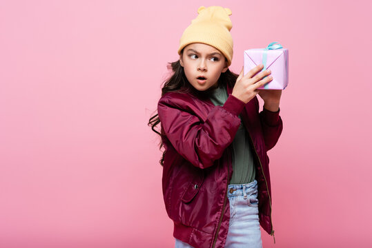 Curious Preteen Kid In Trendy Outfit Shaking Wrapped Present Isolated On Pink.